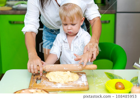 happy little boy preparing dough in kitchen at table. there are dough products on table, dressed as chef happy little boy preparing dough in kitchen at table. there are dough products on table, dressed as chef 108485953