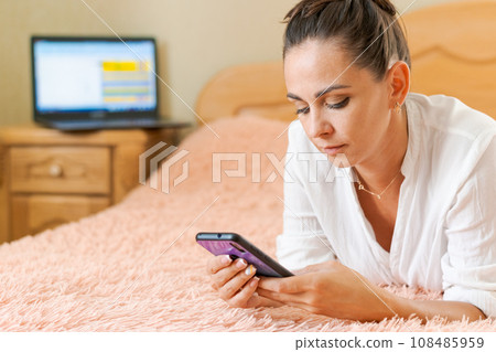 cute woman lying on the bed with a phone in her hand on a soft pink blanket 108485959