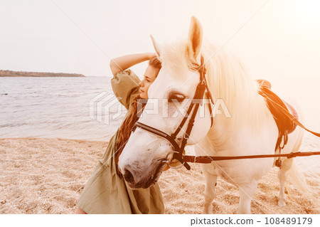 A woman in a dress stands next to a white horse on a beach, with the blue sky and sea in the background. A woman in a dress stands next to a white horse on a beach, with the blue sky and sea in the background. 108489179