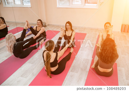 A group of six athletic women doing pilates or yoga on pink mats in front of a window in a beige loft studio interior. Teamwork, good mood and healthy lifestyle concept. A group of six athletic women doing pilates or yoga on pink mats in front of a window in a beige loft studio interior. Teamwork, good mood and healthy lifestyle concept. 108489203