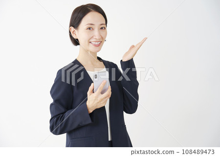 中年女商人拿著智慧型手機，用手掌、白色背景指著右側 108489473