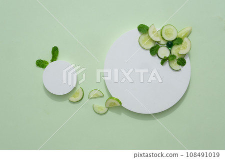 Empty cylindrical platform for cosmetics with cucumber ingredient. Mockup with concrete circle decorated with cucumber slices and mint leaves on green background Empty cylindrical platform for cosmetics with cucumber ingredient. Mockup with concrete circle decorated with cucumber slices and mint leaves on green background 108491019
