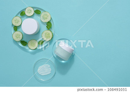 Against the light blue background, cosmetic jars without label decorated with fresh slices of cucumber and mint leaves on transparent podiums. Mockup for design, advertising photo for cosmetic Against the light blue background, cosmetic jars without label decorated with fresh slices of cucumber and mint leaves on transparent podiums. Mockup for design, advertising photo for cosmetic 108491031