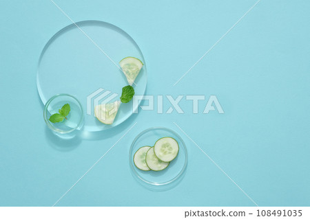 On a pastel blue background, petri dishes and transparent podiums containing slices of cucumber and mint leaves. Blank space for display product and design. Top view, natural ingredient for skin care On a pastel blue background, petri dishes and transparent podiums containing slices of cucumber and mint leaves. Blank space for display product and design. Top view, natural ingredient for skin care 108491035