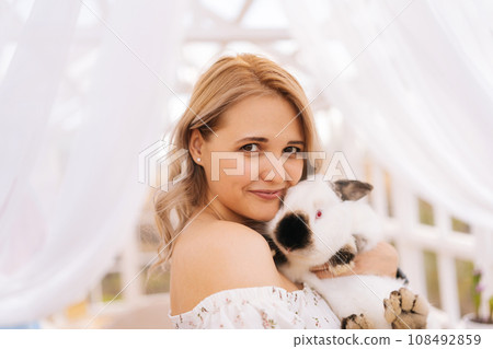 Close-up portrait of charming blonde female in summer dress cuddling adorable fluffy white bunny standing inside of gazebo house, smiling looking at camera. Concept of excursion to eco-farm. 108492859