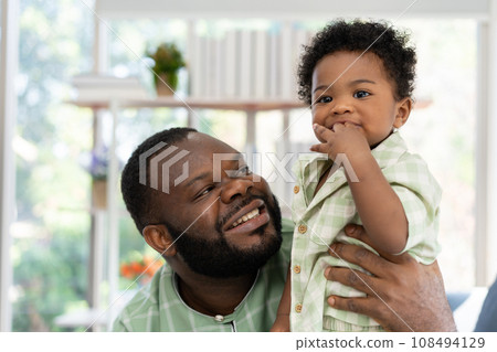 Happy african american man play with little infant toddler child in living room sofa. Parent and little kid relaxing at home. Parents and toddler child girl enjoying spending time at home. Childcare 108494129