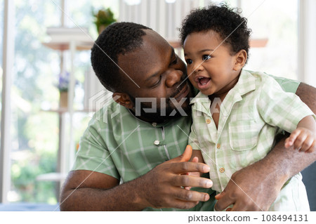 Happy african american man play with little infant toddler child in living room sofa. Parent and little kid relaxing at home. Parents and toddler child girl enjoying spending time at home. Childcare 108494131