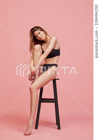 Beautiful young woman with slim fit body posing in underwear, sitting on chai against pink studio background. Body care Beautiful young woman with slim fit body posing in underwear, sitting on chai against pink studio background. Body care 108494200