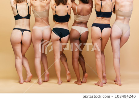 Back view of female bodies of different shapes and skin color standing in underwear against beige studio background. Buttocks and legs 108494240