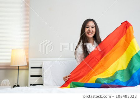 Portraits of happy Asian lesbian girls sitting with rainbow flags on sitting bed beside the window. Cheerful young adult women living with relationship concept. LGBTQ Lifestyle Pride Month 108495328