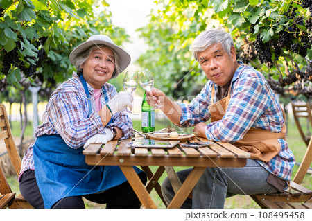 Portrait of senior winemaker holding in his hand a glass of new white wine. Smiling happy elderly couple enjoying a picnic together in own vineyard. Agricultural concept, Small business, retirement Portrait of senior winemaker holding in his hand a glass of new white wine. Smiling happy elderly couple enjoying a picnic together in own vineyard. Agricultural concept, Small business, retirement 108495458