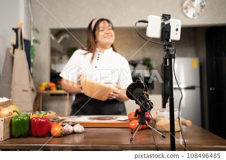Young attractive Asian woman is blogging for her kitchen channel about homemade pizza while streaming online for webinar masterclass lesson. Vlogger broadcasting live video online teaching cooking 108496485