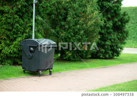 Large trash container in a city park. Green plastic trash can standing near trees 108497338