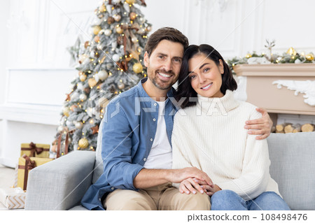 Portrait of young loving family couple, man and woman hugging and looking at camera, sitting on sofa in living room cinema of apartment, celebrating new year and christmas together. Portrait of young loving family couple, man and woman hugging and looking at camera, sitting on sofa in living room cinema of apartment, celebrating new year and christmas together. 108498476