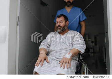 Male nurse pushing a patient in a wheelchair along a hospital corridor. Overweight patient feeling anxious and has health concerns. 108498485