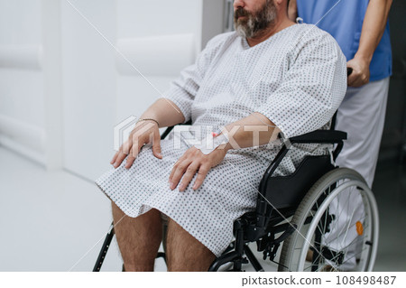 Male nurse pushing a patient in a wheelchair along a hospital corridor, waiting for medical examination. Overweight patient feeling anxious and has health concerns. 108498487