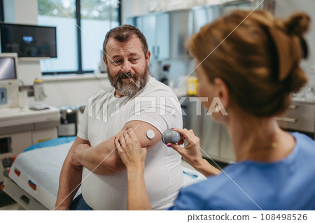 Doctor applying a continuous glucose monitor sensor on patient's arm. Obese, overweight man is at risk of developing type 2 diabetes. Concept of health risks of overwight and obesity. 108498526