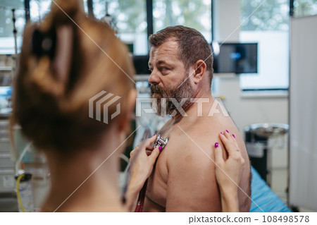 Female doctor listening to heartbeats and rhytm of overweight patient using stethoscope, detecting murmurs, irregularities, heart palpitations. Obesity affecting middle-aged men's health. Concept of Female doctor listening to heartbeats and rhytm of overweight patient using stethoscope, detecting murmurs, irregularities, heart palpitations. Obesity affecting middle-aged men's health. Concept of 108498578