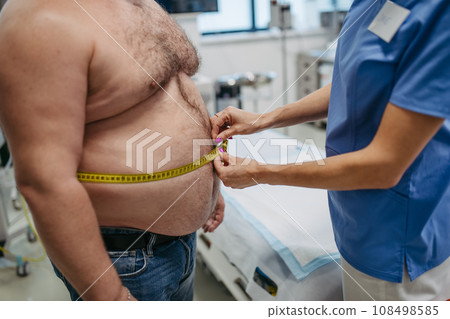 Female doctor measuring waist of overweight patient using tape measure. Obesity affecting middle-aged men's health. Concept of health risks of overwight and obesity. 108498585