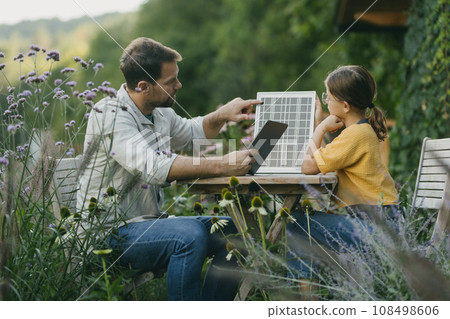 Father teaching daughter about solar panels, photovoltaic, showing her results on tablet. Ssolar energy and sustainable lifestyle of young family. Concept of green energy and sustainable future for Father teaching daughter about solar panels, photovoltaic, showing her results on tablet. Ssolar energy and sustainable lifestyle of young family. Concept of green energy and sustainable future for 108498606