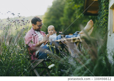 Man sitting in the garden with feet on table, playing with his baby son. Father having bonding moment with his little kid. Baby playing with flower. Man sitting in the garden with feet on table, playing with his baby son. Father having bonding moment with his little kid. Baby playing with flower. 108498612