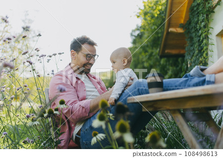 Man sitting in the garden with feet on table, playing with his baby son. Father having bonding moment with his little kid. Man sitting in the garden with feet on table, playing with his baby son. Father having bonding moment with his little kid. 108498613