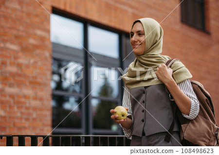 Beautiful woman in hijab standing on city street. Muslim studnet eating apple, going to school. Iran, Afganistan female teacher in front of school building. Beautiful woman in hijab standing on city street. Muslim studnet eating apple, going to school. Iran, Afganistan female teacher in front of school building. 108498623