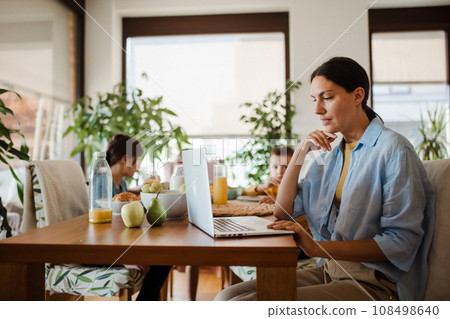 Mother working on laptop at kitchen table while children have breakfast. Remote work and home office for mothers with children. Mother working on laptop at kitchen table while children have breakfast. Remote work and home office for mothers with children. 108498640