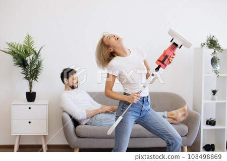 Woman dancing with cordless vacuum while man using laptop Woman dancing with cordless vacuum while man using laptop 108498869