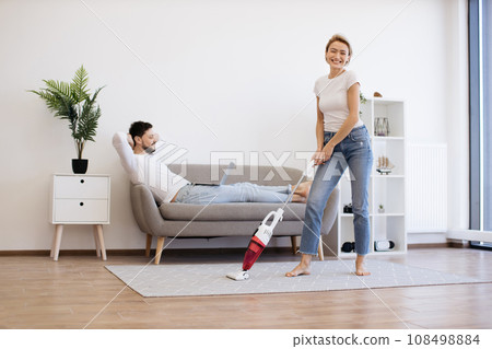 Husband working on laptop while wife vacuuming floor 108498884