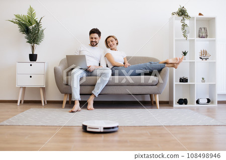 Caucasian couple using laptop while robot cleaning floor 108498946