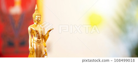 Standing Buddha statue is placed on top of a rectangular object in front of a red framed picture and tree house. Standing Buddha statue is placed on top of a rectangular object in front of a red framed picture and tree house. 108499018