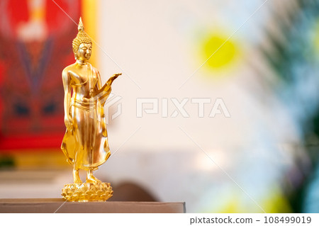 Standing Buddha statue is placed on top of a rectangular object in front of a red framed picture and tree house. Standing Buddha statue is placed on top of a rectangular object in front of a red framed picture and tree house. 108499019