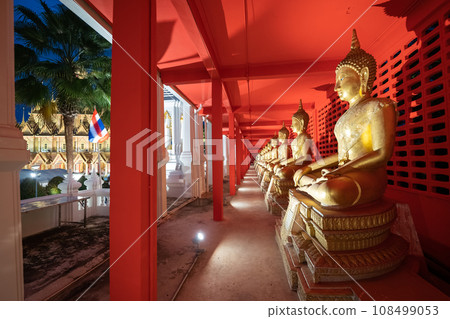 Several golden statues of Buddha statues in meditation posture are lined up in a corridor and spotlight on every statues in front of the red wall. 108499053