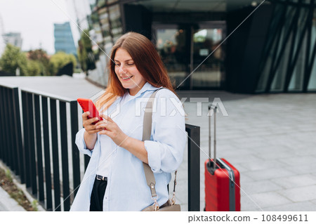 Young woman with a red suitcase using smart phone at airport. Redhead girl in international airport walking with her luggage. Concept of travel Young woman with a red suitcase using smart phone at airport. Redhead girl in international airport walking with her luggage. Concept of travel 108499611