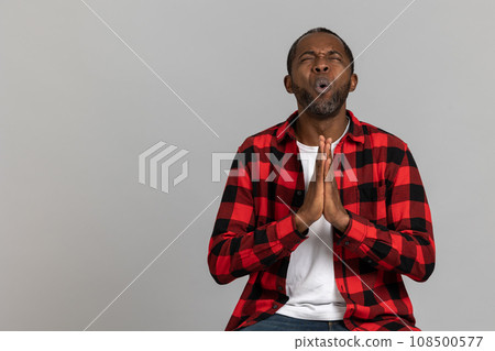Pleading black bearded man wearing red checkered shirt praying for success 108500577