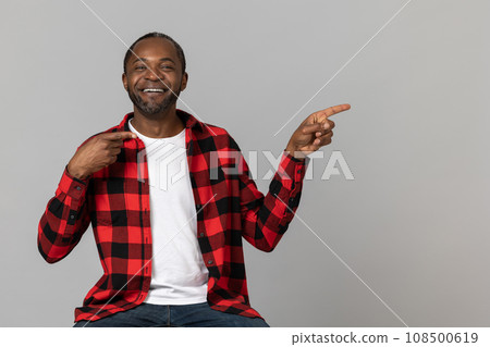 Positive black bearded man wearing red checkered shirt pointing at advertisement area Positive black bearded man wearing red checkered shirt pointing at advertisement area 108500619