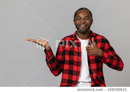 Joyful black bearded man wearing red checkered shirt presenting copy space on palm showing thumb up 108500631