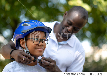 Dark-skinned man helping his kid to put on a protective hemlet Dark-skinned man helping his kid to put on a protective hemlet 108504765
