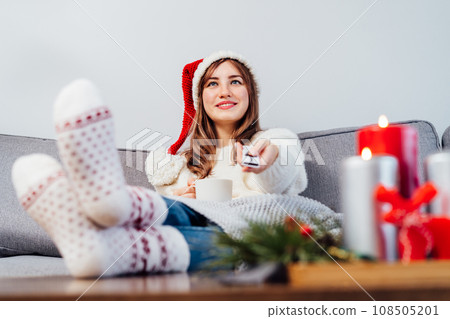 Relaxed, smiling woman in santa hat, jumper and warm socks sitting on comfortable couch, using tv remote controller and watching movie, TV with christmas decoration atmosphere. Cozy winter concept. Relaxed, smiling woman in santa hat, jumper and warm socks sitting on comfortable couch, using tv remote controller and watching movie, TV with christmas decoration atmosphere. Cozy winter concept. 108505201