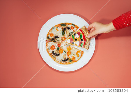 Merry Christmas food offers. Top view hand takes a fake pizza slice on the white plate with veggie pizza on red background. Creative concept for seasonal and festive greetings for pizzeria delivery 108505204