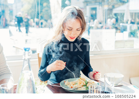 Young Caucasian female sitting at served table in casual cafe and eating delicious hot risotto with shrimps or curry rice during lunch break. Try new foods while traveling. real people. copy space. 108505224