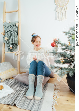 Young woman in cozy sweater with cup of hot drink decorating potted Christmas tree with glass baubles in light modern Scandinavian interior. Eco-friendly winter holidays. Christmas tree in a pot. 108505245