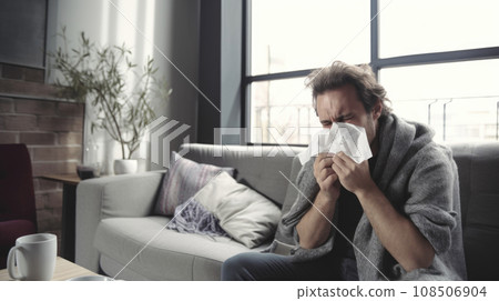 Sick guy with a tissue, sneezing on the sofa Sick guy with a tissue, sneezing on the sofa 108506904