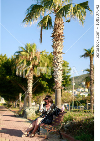 A brunette woman with glasses is sitting on a bench on the street near palm trees 108507642
