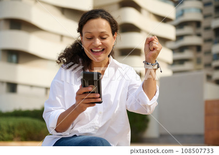 Happy woman clenching fist and rejoicing reading good new on her smartphone, sitting outdoors against high ride building 108507733