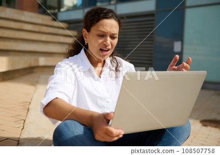 Young female student sitting outdoors on steps with laptop on knees, gesturing with hand, expressing anger failing exams 108507758
