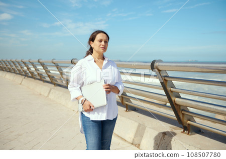 Young woman holding laptop, thoughtfully looking into the distance, strolling the street against seashore background 108507780