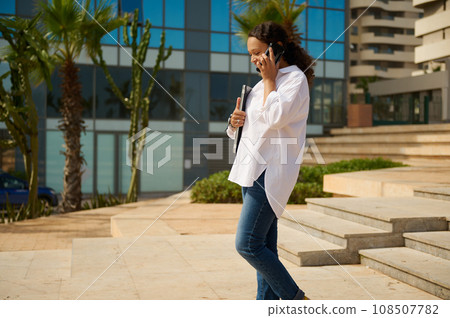 Happy young brunette female manager in casual attire, holding laptop and talking on mobile phone while walking outdoors in city. 108507782