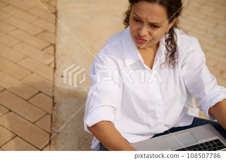 Mixed race young woman expressing sadness failing business tender project, looking aside, sitting with laptop outdoors 108507786